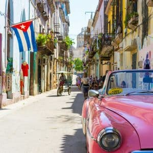 Un coche clásico descapotable rosa está estacionado en una calle soleada de la ciudad, flanqueada por edificios coloridos, con una bandera cubana colgando de uno de ellos.