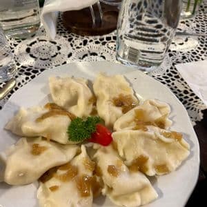 A white plate of pierogi dumplings topped with fried onions, garnished with parsley and tomato, sits on a lace doily.