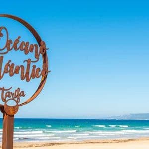 Un cartel metálico oxidado que dice 'Océano Atlántico Tarifa' se encuentra en una playa de arena con el océano turquesa de fondo.