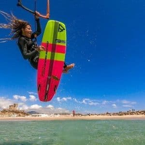 Una mujer en traje de neopreno vuela con su kiteboard sobre el mar en un día soleado, con una playa y un perro de fondo.