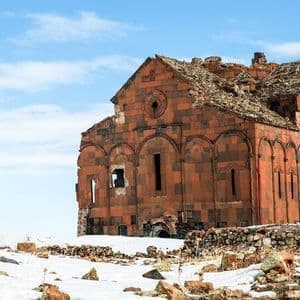 Una cattedrale diroccata in pietra rossa si erge su una collina innevata sotto un cielo azzurro splendente.