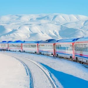 Un treno passeggeri viaggia su un binario curvo attraverso un paesaggio innevato con montagne sotto un cielo azzurro.