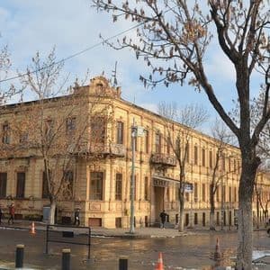Un grande edificio storico giallo pallido all'angolo di una strada, con alberi spogli e persone che camminano sul marciapiede bagnato.