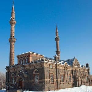 Una moschea in pietra riccamente decorata con due minareti si erge in un paesaggio innevato sotto un cielo azzurro e limpido, con alberi brinati a sinistra.