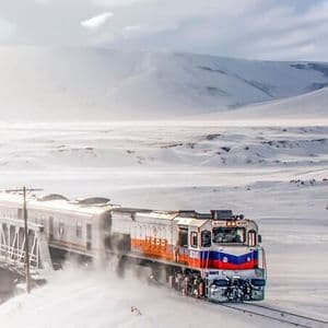 Un treno con una locomotiva rossa e blu attraversa un paesaggio innevato, sollevando neve mentre si muove.
