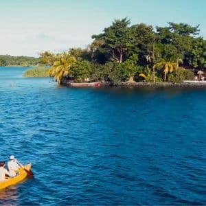 Two people in a yellow kayak paddling across a blue lake towards a small, tree-covered island.