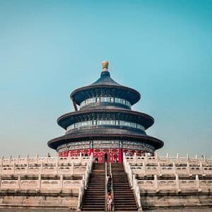 Una vista dal basso di un tradizionale tempio cinese a tre livelli con tetto blu, in cima a una larga scalinata in pietra sotto un cielo azzurro e limpido.