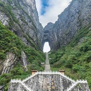 Una lunga scalinata ripida sale su una montagna ricoperta di verde verso un imponente arco di roccia naturale situato tra due scogliere.