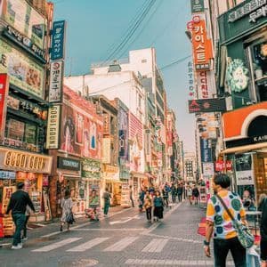 Pedoni su una trafficata strada cittadina, che camminano tra edifici coperti di insegne colorate in caratteri coreani sotto un cielo sereno.