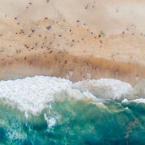 Vista aerea dall'alto di una spiaggia sabbiosa affollata con persone sparse lungo la riva mentre onde turchesi si infrangono.