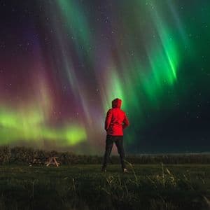 Una persona con una chaqueta roja con capucha está de pie en un campo de hierba por la noche, observando la aurora boreal verde y morada en el cielo estrellado.