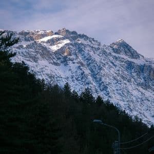 La luce del sole illumina le cime di una grande montagna innevata che sovrasta una fitta e scura foresta di pini sotto un cielo blu.