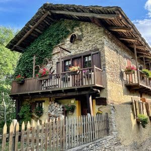 Una casa rustica in pietra con muri coperti di edera, balconi in legno fioriti e una staccionata in un ambiente rurale.