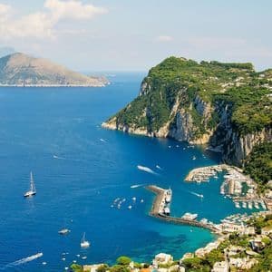 Ein erhöhter Blick auf den Yachthafen einer Küstenstadt, gefüllt mit Booten, eingebettet neben einer grünen, felsigen Klippe unter blauem Himmel.