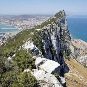 Una vista elevada desde una cresta rocosa de montaña, dominando una ciudad costera, un puerto y el mar azul profundo en un día soleado.