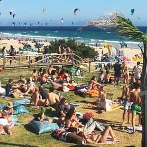 Un gran grupo de personas tomando el sol en un césped con vistas a una playa llena de kitesurfistas bajo un cielo azul claro.