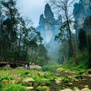 Un viaggio di gruppo WeRoad si trova accanto a un ruscello e un ponte di legno in una foresta, con imponenti formazioni rocciose avvolte nella nebbia sullo sfondo.