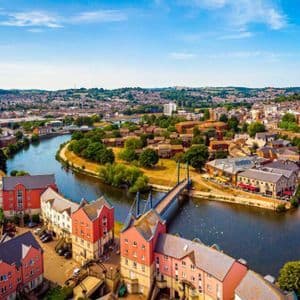 Veduta aerea di una città su un fiume sinuoso, con edifici colorati sul lungofiume, un ponte e colline verdi sotto un cielo azzurro.