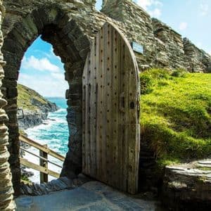 Una porta di legno aperta in un arco di pietra rivela una vista dell'oceano turchese che si infrange contro una costa rocciosa.