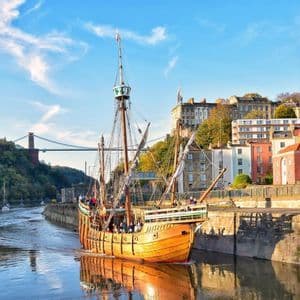 Una storica nave di legno con passeggeri a bordo naviga lungo un fiume, con uno skyline cittadino e un grande ponte sospeso sullo sfondo.
