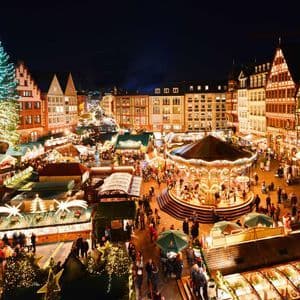 An elevated view of a bustling Christmas market at night, with a large illuminated tree, a glowing carousel, and market stalls.