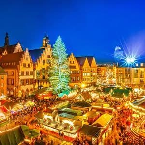 An elevated view of a bustling Christmas market in a city square at night, with a large illuminated tree, a carousel, and festive stalls.