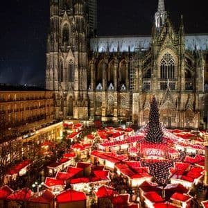 An elevated view of a bustling Christmas market at night, with red-roofed stalls and a large tree, set against an illuminated Gothic cathedral.