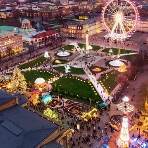An aerial view of a bustling city square at dusk, filled with a Christmas market, an illuminated Ferris wheel, and an ice rink.