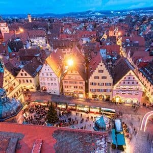 An aerial view of a historic European town square bustling with a Christmas market at twilight, with lights illuminating the gabled buildings.