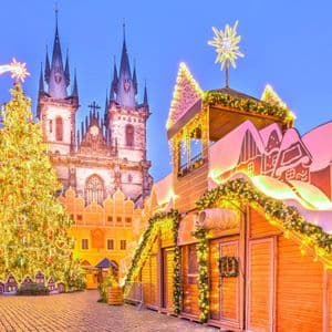 A brightly lit Christmas tree and market stalls fill a historic cobblestone square in front of a Gothic church at dusk.
