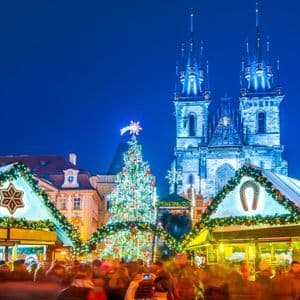 A crowded Christmas market at night with an illuminated tree and stalls in front of a large Gothic church.
