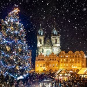 A large, illuminated Christmas tree stands in a bustling town square during a snowy Christmas market at night.