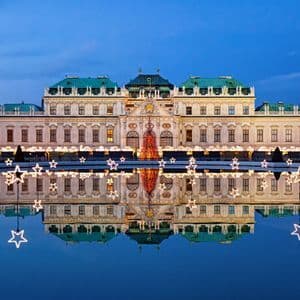 An ornate palace illuminated for Christmas with a large lighted tree and stars, reflected perfectly in a pool of water at dusk.