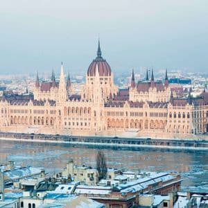 A grand, ornate building with a central dome sits on the banks of an icy river, with snow-covered city rooftops in the foreground.