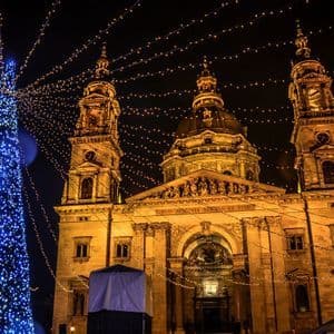 An illuminated basilica at night, with a large Christmas tree covered in blue lights and festive string lights overhead.