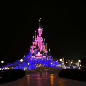 Ein großes Schloss, beleuchtet mit rosa und blauen Lichtern vor einem dunklen Nachthimmel, während eine Person auf dem nassen Weg darauf zugeht.