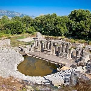 The ancient ruins of a stone amphitheater with a flooded stage, surrounded by green trees and mountains under a clear blue sky.