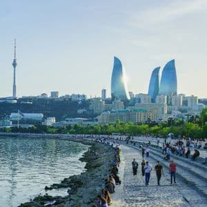 Persone che si rilassano su una passeggiata lungomare con uno skyline moderno che presenta torri a forma di fiamma e una grande torre televisiva.