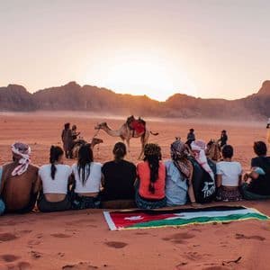 Un groupe WeRoad assis sur une dune de sable rouge, avec un drapeau jordanien, admirant le coucher de soleil sur le désert, avec des montagnes et des chameaux en arrière-plan.