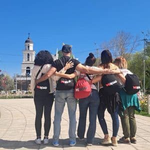 Un gruppo WeRoad di sei persone, abbracciate, di fronte a un campanile in un parco in una giornata di sole.