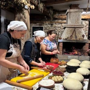 Tre donne preparano dolci e impasti su un lungo tavolo all'interno di una cucina rustica con un grande camino in pietra.