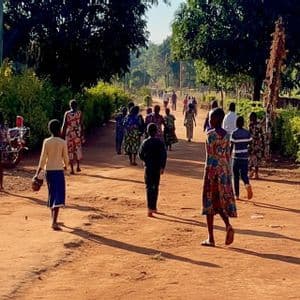 Un gruppo di adulti e bambini si allontana dalla fotocamera su un ampio sentiero sterrato, fiancheggiato da una vegetazione lussureggiante e alberi.
