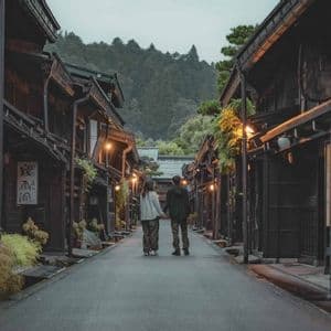 Una coppia si tiene per mano, vista da dietro, in una stretta via fiancheggiata da tradizionali edifici giapponesi in legno, al crepuscolo.