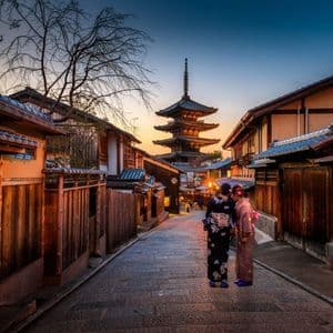 Due donne in kimono tradizionali su una strada acciottolata osservano un'alta pagoda al tramonto.