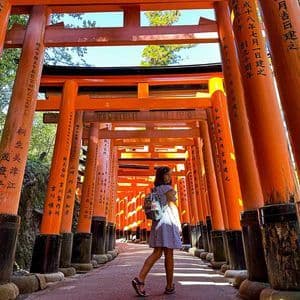Una donna con uno zaino cammina lungo un sentiero costeggiato da numerosi torii arancioni, voltandosi indietro.