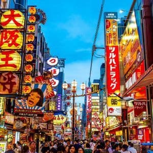Una strada affollata in una città giapponese al tramonto, costeggiata da edifici coperti di insegne e lanterne colorate e illuminate.