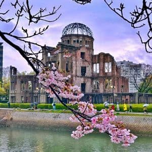 Rami di ciliegio in fiore rosa incorniciano la vista di un edificio in rovina con una cupola scheletrica attraverso un fiume in un parco cittadino.