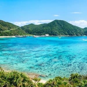 Vista panoramica di una baia tropicale con spiaggia sabbiosa, acqua turchese cristallina e rigogliose colline verdi sotto un cielo azzurro.