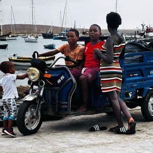 Quattro bambini si radunano attorno a un triciclo motorizzato blu su un molo, con un porto pieno di barche sullo sfondo.