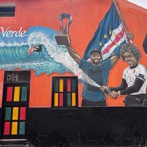 Un murale colorato su un edificio arancione raffigura tre atleti che celebrano una vittoria negli sport acquatici con un trofeo e la bandiera di Capo Verde.